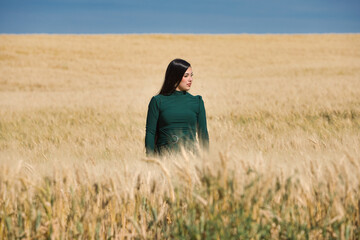 A beautiful, dark-haired teenage Latin girl dressed in green stands among a dry, yellow wheat field on a summer day. In the background, the blue sky.