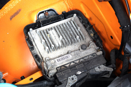 Close-up of a dusty van engine control unit ECU with cooling fins and wiring, mounted on an orange metal firewall inside a vehicle engine bay. Car engine control module ECM.