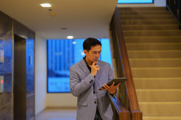 Young businessman is thinking while using a tablet in a modern office building, standing near the staircase and elevator, reflecting the challenges and opportunities of corporate life