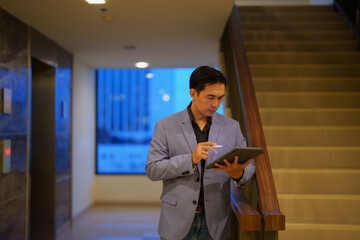 Young manager working with digital tablet and stylus pen, standing in the corridor of a modern office building near the elevator and the stairs