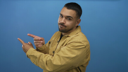 Hispanic man wearing tan jacket and black shirt pointing finger to side in studio against blue wall; confidence.