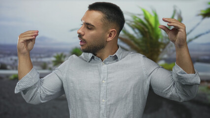 Young hispanic man talking with hands by palm trees in front of building outdoors wearing striped shirt with rolled sleeves; communication connection energy enthusiasm.