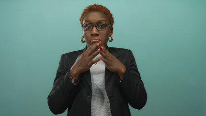 Woman wearing glasses and hoop earrings covers mouth with hands against teal studio background;...