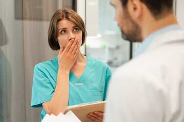 Shocked nurse covering mouth discussing with doctor in hospital corridor