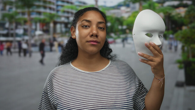 African american woman holding a white plaster mask to her face in a busy urban street with blurred passersby; mystery.