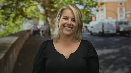 Woman smiling with bare shoulder in front of a city street with trees and parked cars, wearing a black top; happiness.