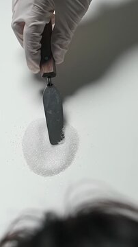 Pristine white-gloved hand meticulously levels luminous white granular material with a miniature silver precision spatula on an immaculate white background, captured in an extreme close-up,
