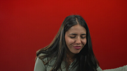 Young woman strums air guitar with hands against red studio wall while smiling and leaning forward; playful joy.