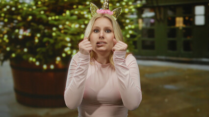 Woman outdoors in city wearing unicorn diadem looks nervous with hands raised, blending humor and anxiety against backdrop of blurred lights in the street setting.