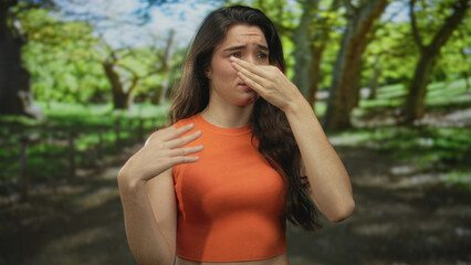 Young hispanic woman wearing an orange sleeveless top in green park pinches her nose to block a...