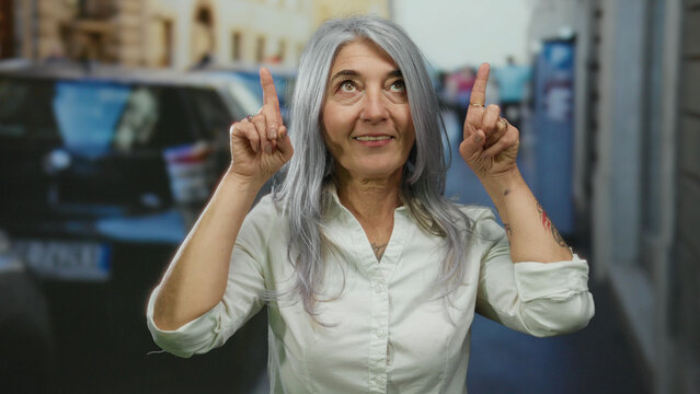 Senior woman with grey hair outdoors on a street joyfully raising her fingers as if pointing at something inspiring above the city landscape.