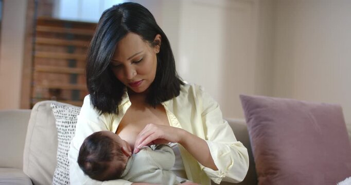 Mother unbuttoning shirt, exposing breast and cradling infant for feeding on sofa in living room
