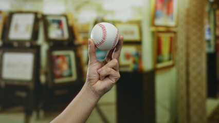 Caucasian man's hand holding a baseball in a brightly lit museum with blurred art in the background, highlighting the focus on the ball and the indoor setting.
