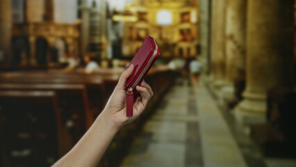 Hand holding red wallet indoors in church setting with blurred background featuring church pews and columns under soft lighting, capturing elegant simplicity and atmosphere.