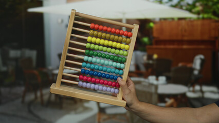 Man holding colorful abacus on outdoor restaurant terrace with blurred background showcasing tables and chairs, evoking a playful educational scene in a city setting.