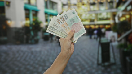 Male hand holding czech koruna banknotes outdoors in a busy city street, indicating commerce and currency exchange in an urban setting.