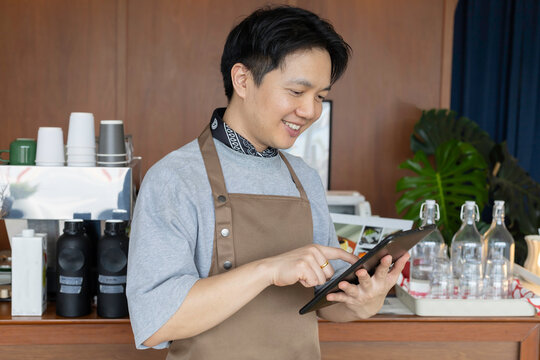Happy Asian barista, a small business owner, using a tablet for orders in his modern cafe. Coffee shop entrepreneur managing operations with technology.