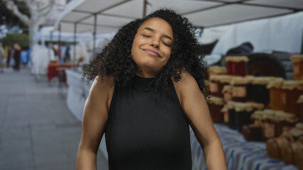 Woman smiles and stretches arms behind head beside stacked jam jars on sunlit street market stall; carefree happiness.