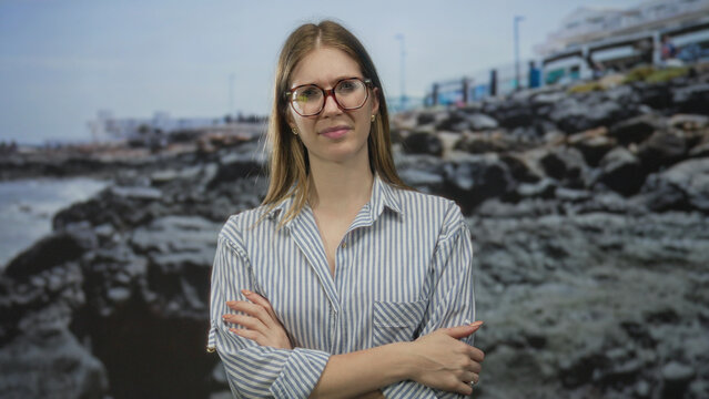 Blonde woman wearing glasses and striped shirt with hand on cheek by rocky seaside beach; doubt concern thought.