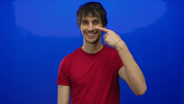 Man pointing finger to nose with broad friendly grin and joyful expression in blue studio; happiness.