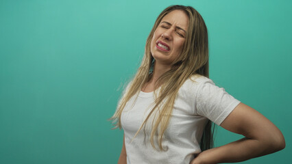 Fototapeta premium Woman clutching lower back in studio with teal background, leaning sideways and wincing in pain; discomfort tension stress.