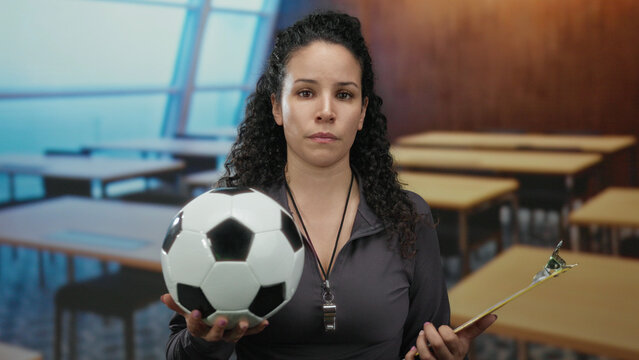 Hispanic woman holding soccer ball and clipboard in classroom with focused expression, teaching sports indoors in educational setting, wearing whistle and dark jacket