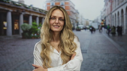 Young woman with long blonde hair and glasses stands confidently on a bustling urban street with historic architecture in the background.