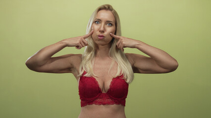 Blonde woman pinches cheeks with fingers in green studio setting while looking at camera; joy fun spontaneity playfulness.