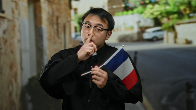 Young asian priest holding french flag on urban street, wearing glasses and clerical attire, with blurred town background highlighting cross necklace and serene expression.