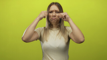 Young blonde woman makes a humorous crying gesture against an isolated yellow background wall, showcasing a playful and exaggerated expression with closed fists near her face.