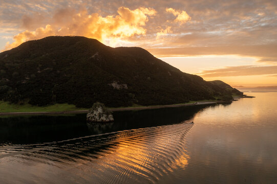 Aerial view of a dramatic sunset casting golden light over calm waters near a lush, dark green headland, creating a striking contrast, Whangarei, Northland Region, New Zealand.