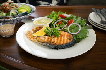 Grilled salmon steak served with fresh green vegetables and salad dressing on wooden table. Represents healthy eating with clean food, a balanced diet, nutritious meal and gourmet seafood cuisine.