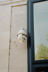 A security camera is fixed on a wall next to a large glass panel, observing the outdoor environment. The setting is bright, indicating daytime, with nearby trees visible.