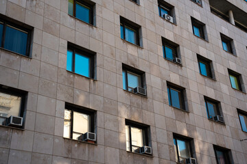 A city block features modern architecture with multiple windows reflecting the vibrant urban environment. Various air conditioning units indicate practical living solutions.