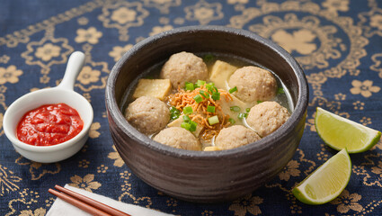 Bakso served in a traditional clay bowl with chili sauce and lime on the side, on a batik tablecloth, top-down view, food styling photography