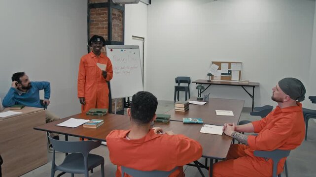 Full shot of young African American male prison inmate in orange jumpsuit standing by whiteboard in classroom, reading essay in front of diverse companions and teacher, who are listening and clapping
