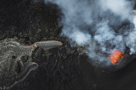 Aerial view of fiery orange lava erupting against the stark black volcanic rock, plumes of white smoke rising, Grindavik, Southern Peninsula Region, Iceland.