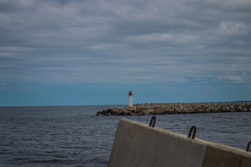 A distant red-topped white lighthouse stands on a breakwater made of concrete tetrapods, under a cloudy sky by the open sea.