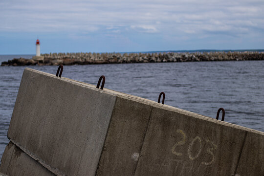 Coastal scene with concrete barrier in foreground, distant lighthouse, and breakwater stretching into a moody sea under cloudy skies.