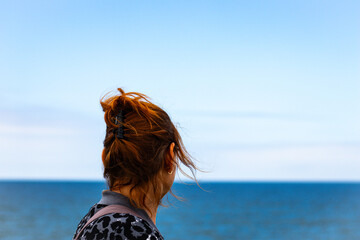 A woman with red hair and a patterned jacket gazes at the ocean horizon, standing against a clear...