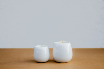 Two white ceramic candles on a brown surface with a light grey background. Minimalistic composition, soft natural lighting. Cozy and peaceful atmosphere for home decor or aromatherapy theme.