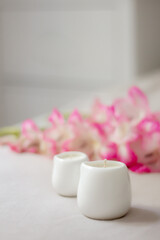 White ceramic candles on a soft surface with blurred pink flowers in the background. Minimalist spa composition for relaxation, wellness, aromatherapy, and home decor concepts.