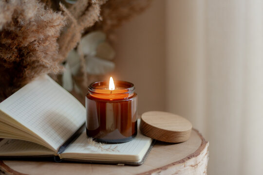 Burning candle in amber glass jar on an open notebook with pen, placed on wooden surface with dried flowers in background. Cozy rustic atmosphere for journaling, inspiration, and relaxation.