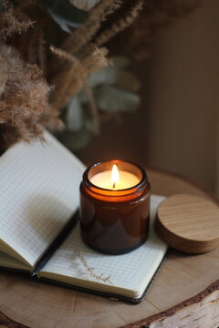 Burning candle in amber glass jar on an open notebook with pen, placed on wooden surface with dried flowers in background. Cozy rustic atmosphere for journaling, inspiration, and relaxation.