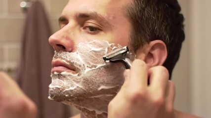 A man is shaving his mustache and beard with a razor in a closed room.