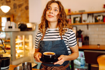 A young female barista stands behind a bar counter with prepared coffee. A beautiful woman with a digital tablet enjoys the sunlight in a cozy coffee shop. Small business, beverage concept.