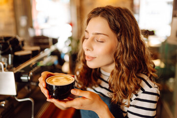 A young female barista stands behind a bar counter with prepared coffee. A beautiful woman with a digital tablet enjoys the sunlight in a cozy coffee shop. Small business, beverage concept.
