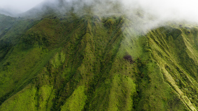 Aerial view of a verdant mountain peak shrouded in mist, its steep slopes etched with deep ravines and lush vegetation, Crater Mont Pelee, Martinique, France.