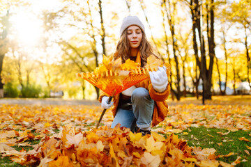 A young woman in a bright jacket and gloves collects bright autumn leaves in a sunny park. A female volunteer collects fallen leaves with a rake. The concept of ecology, cleaning.