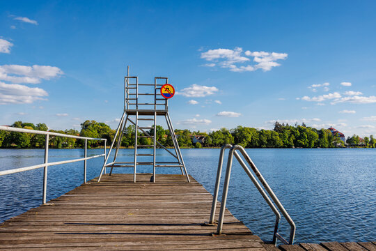 Wooden pier with metal railing and lifeguard tower on the shore of a summer lake. No diving sign on the lifeguard tower. - Powered by Adobe